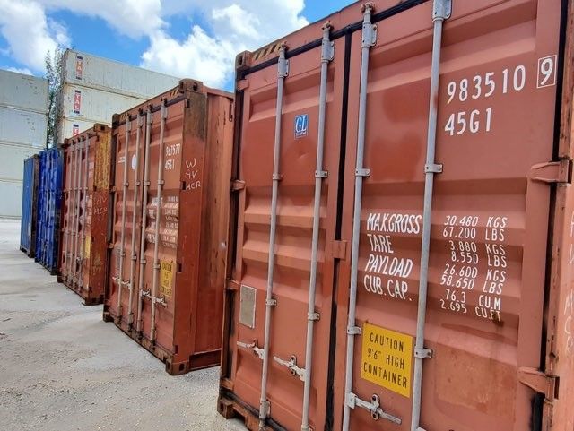 Shipping containers lined up outdoors, various colors including red, brown, and blue, under a blue sky.