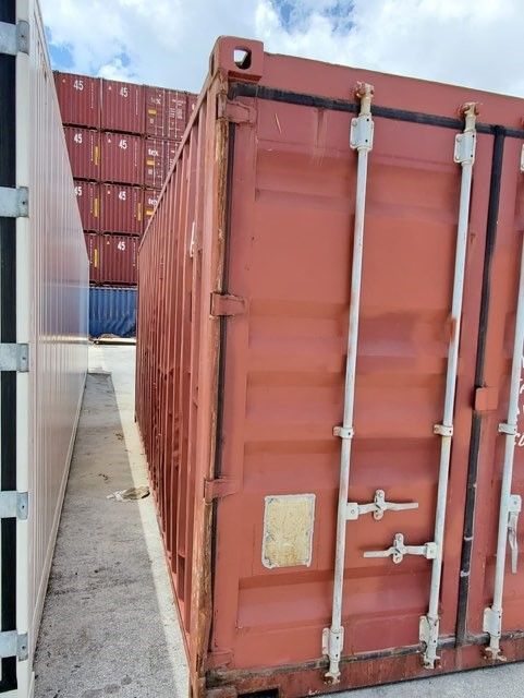 Red shipping container with white latches, standing between a white structure and stacked containers.