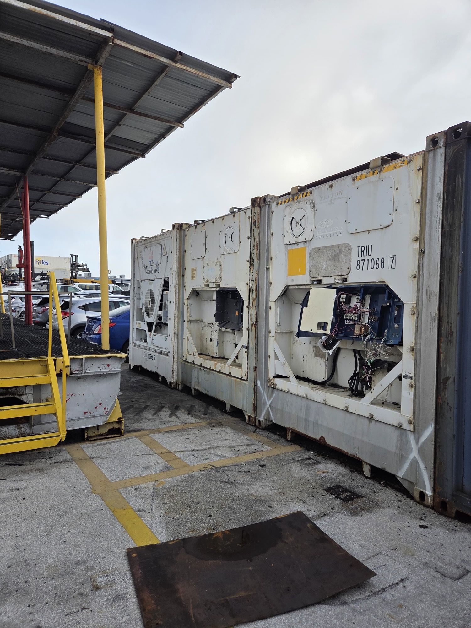 Three white refrigerated shipping containers parked outdoors, next to a covered loading area.