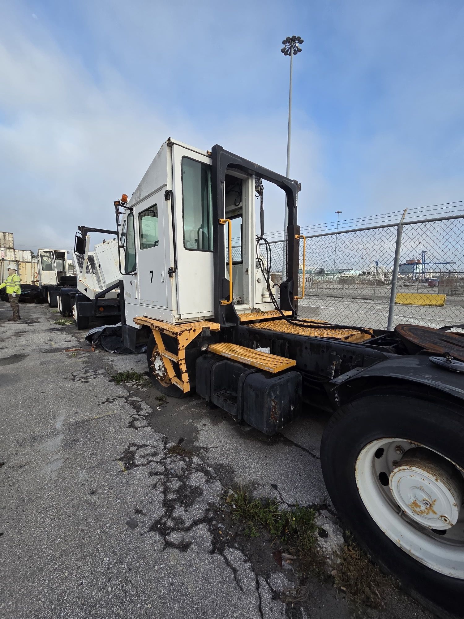 White terminal tractor parked near a chain-link fence on a gray surface with a cloudy sky.