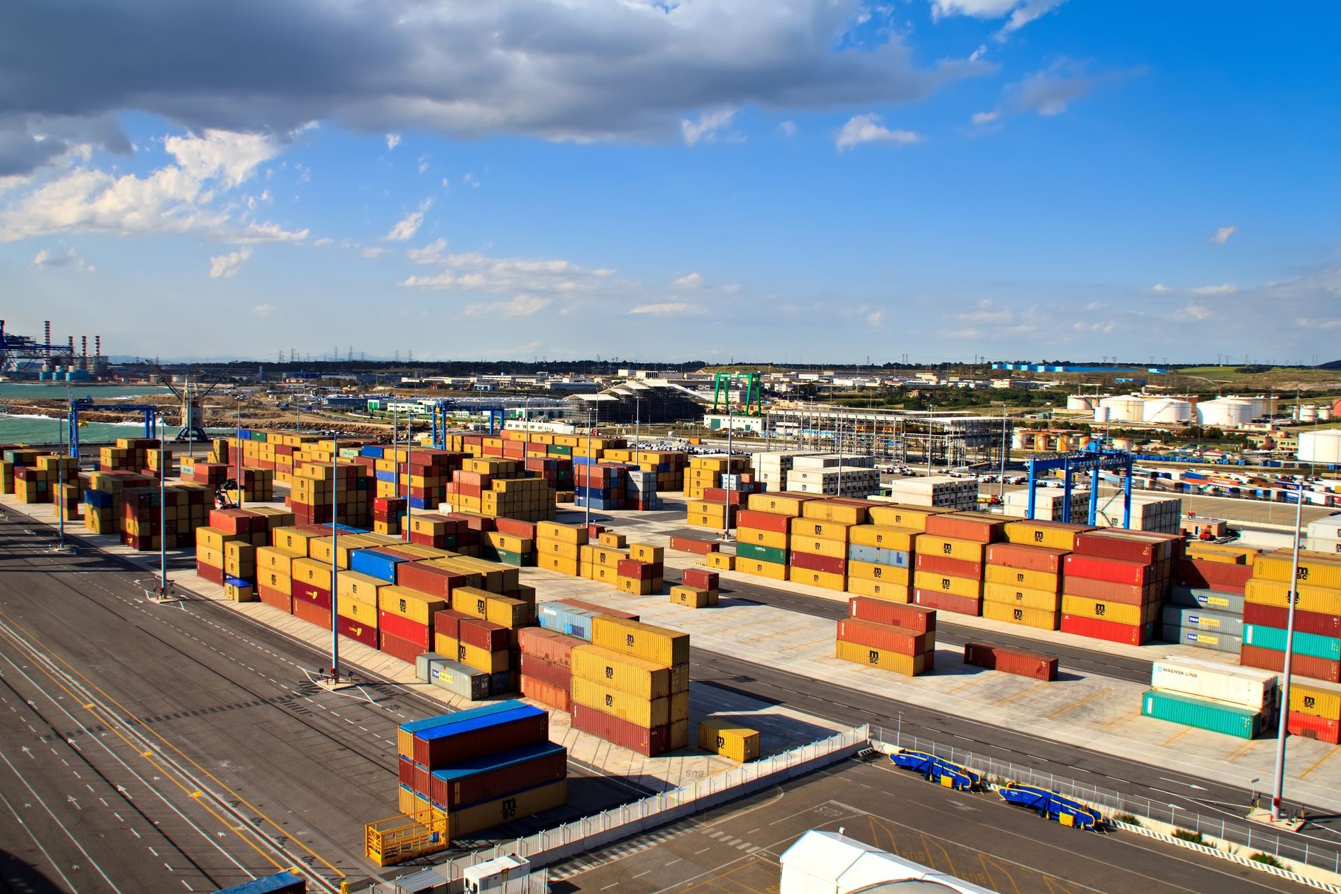 Shipping containers stacked in a port. Yellow, orange, and blue containers sit on a paved area under a blue sky.