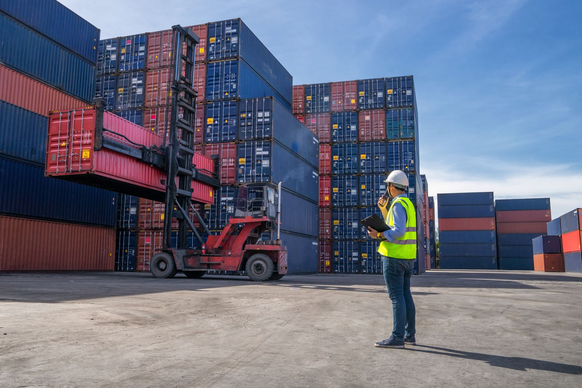 Forklift lifting shipping container in a dockyard, worker in safety vest observing.