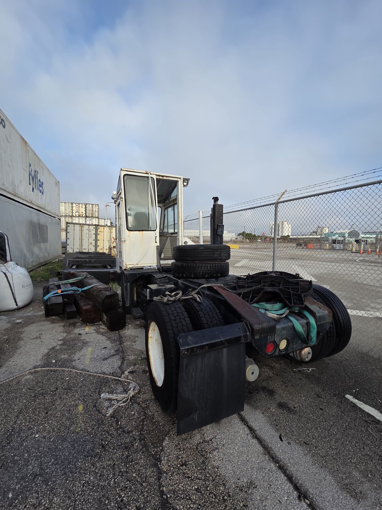 White yard truck, parked outdoors, with stacked tires and shipping containers in the background.