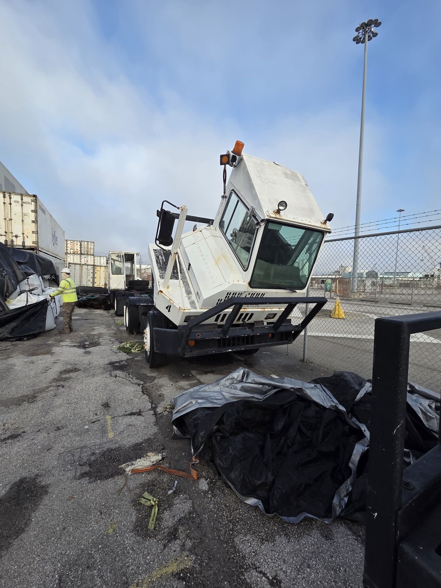 White truck with tilted cab on a wet, paved lot near shipping containers. A person in safety gear stands near.