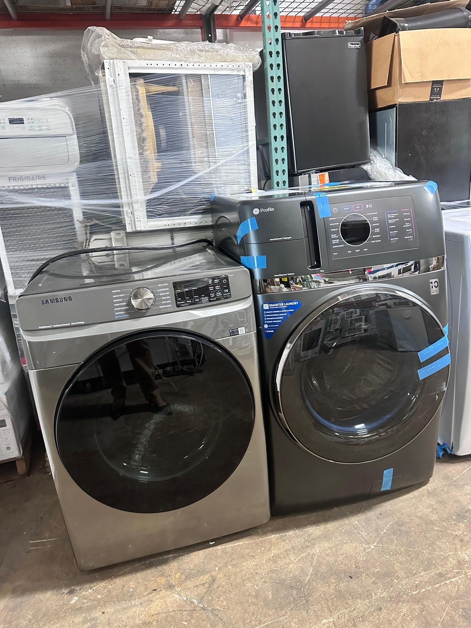 Two washing machines, silver and black, in a warehouse setting.