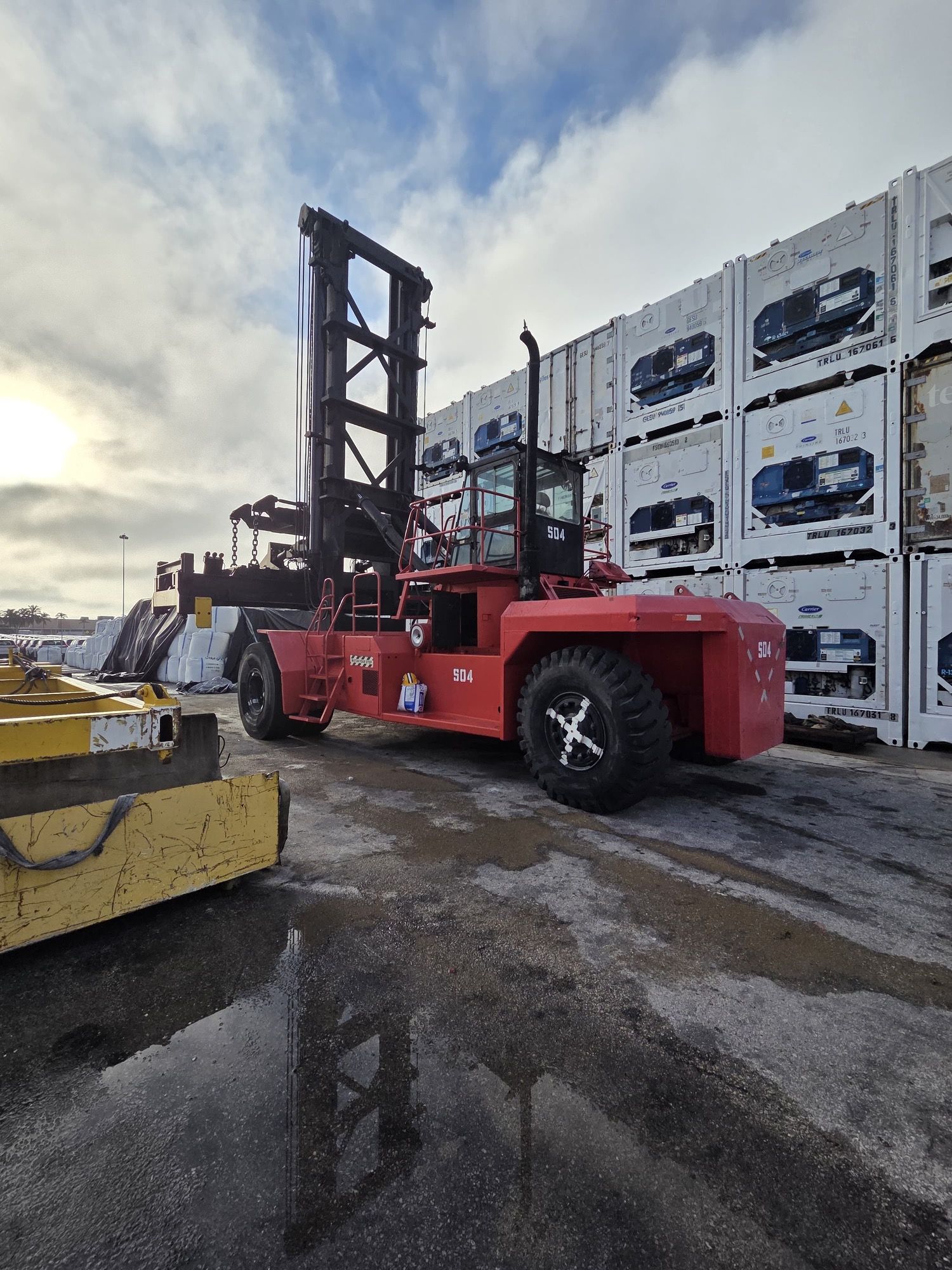 Red container handler at a port, stacking shipping containers. Overcast sky.