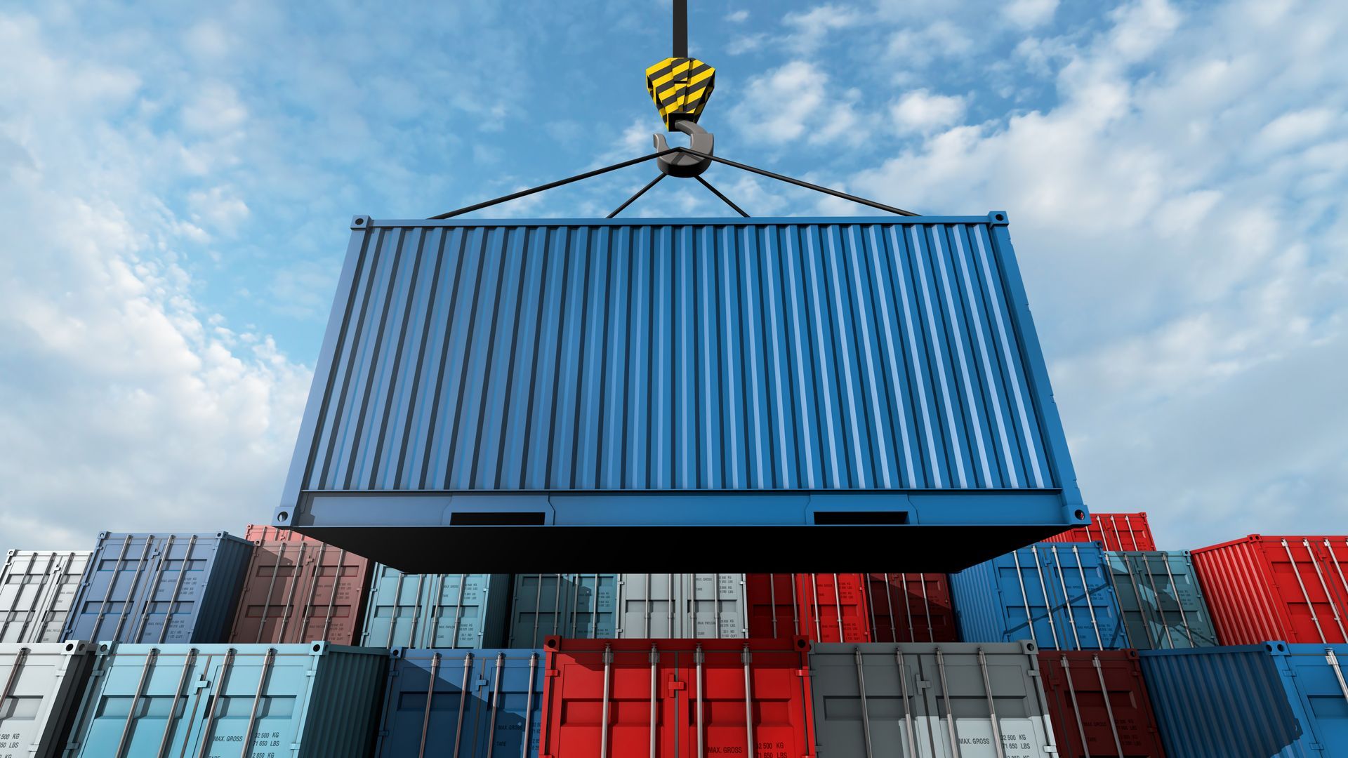 Blue shipping container lifted by a crane above a stack of other colorful containers under a cloudy sky.