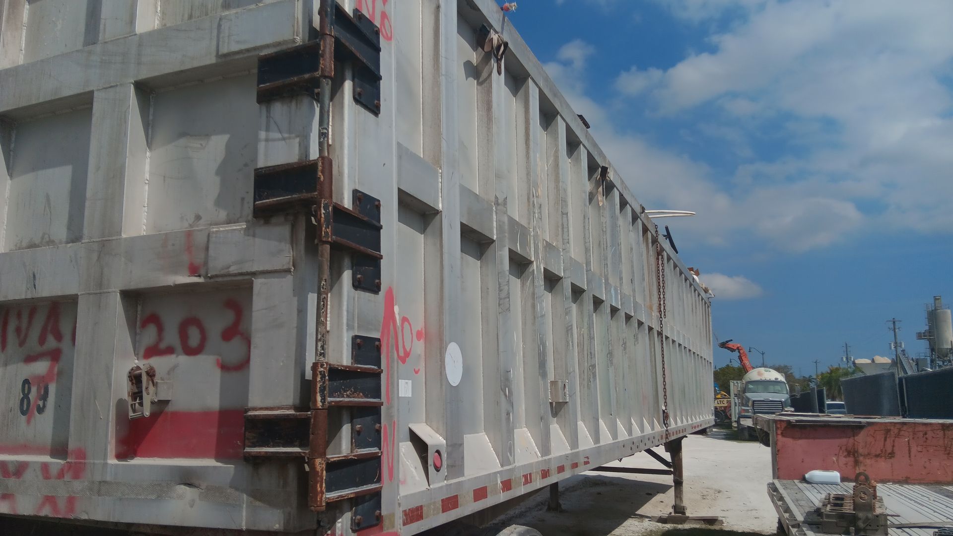 Side view of a large, silver truck trailer with black hinges, parked outdoors on a sunny day.