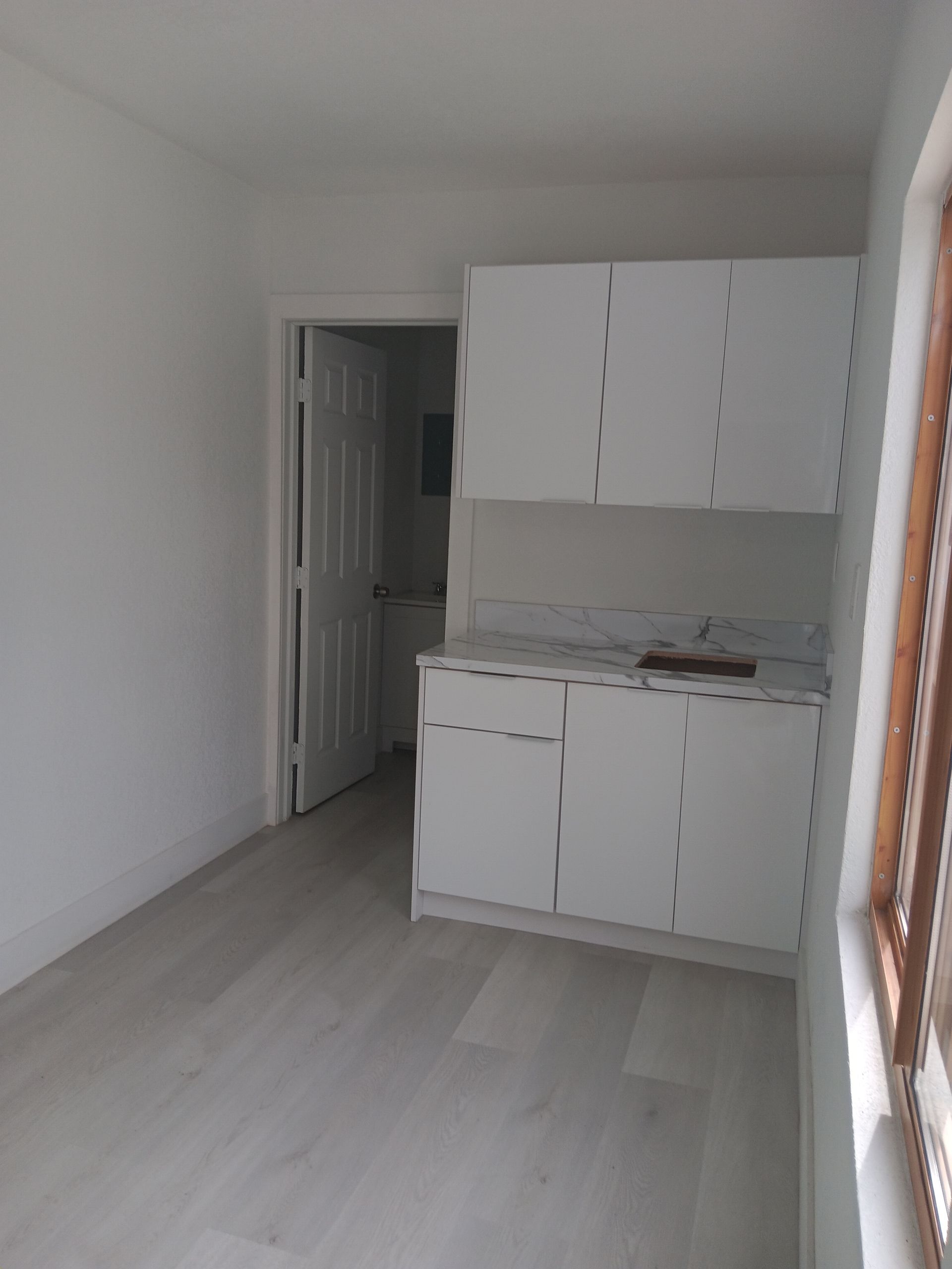 Small white kitchen with cabinets, a window, and a doorway leading to a room.