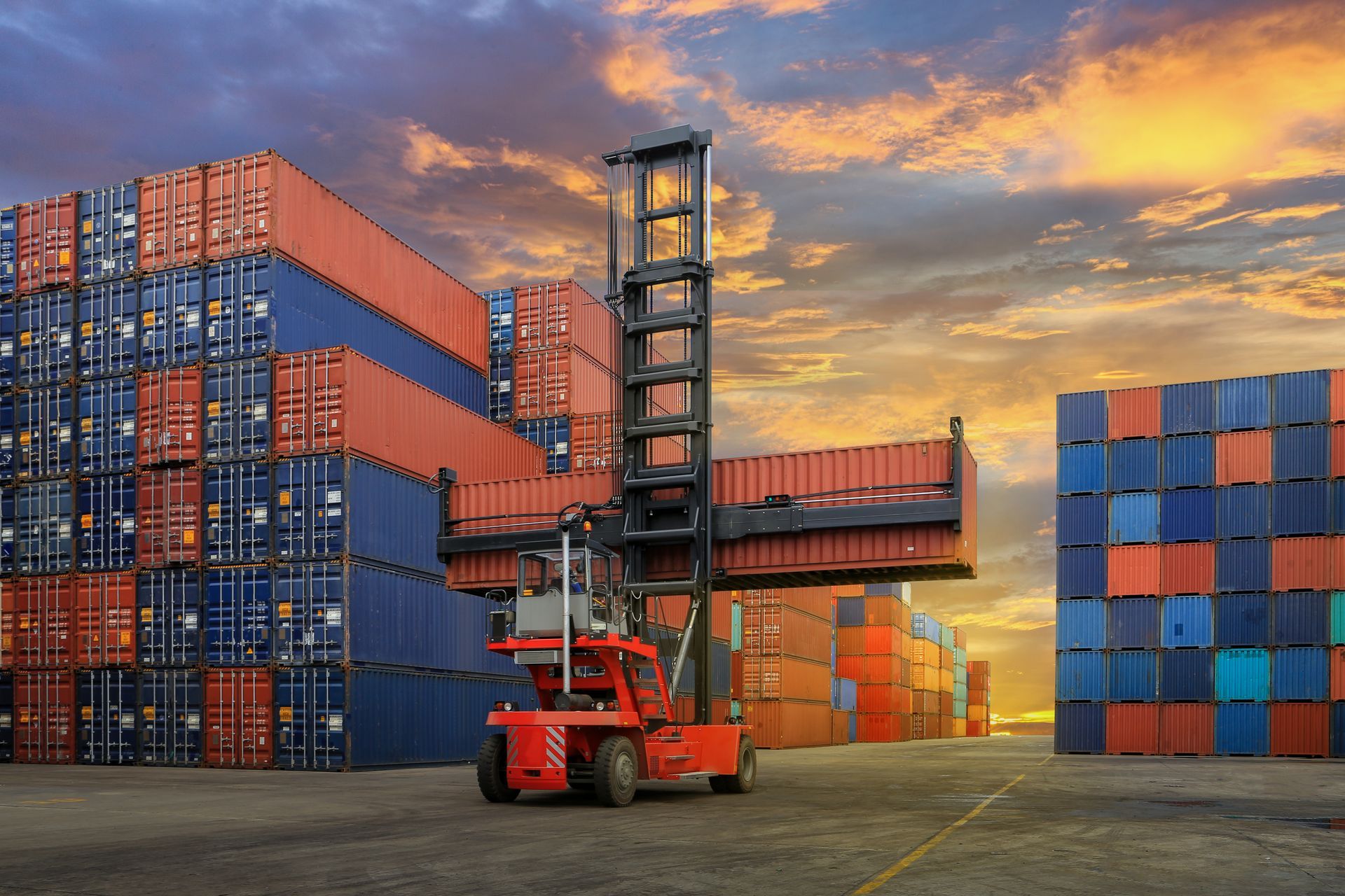 Red forklift lifting a shipping container in a busy port at sunset; stacks of blue and red containers.