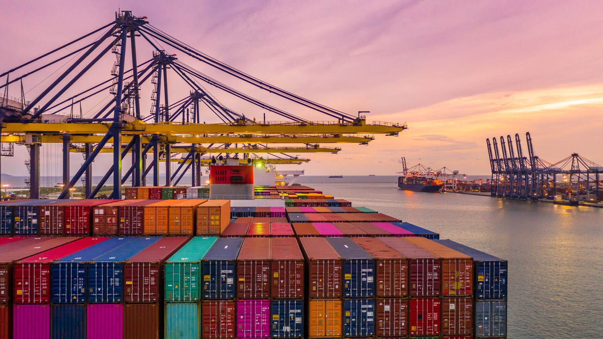 Cargo containers on a ship at a port, with cranes in the background under a sunset sky.
