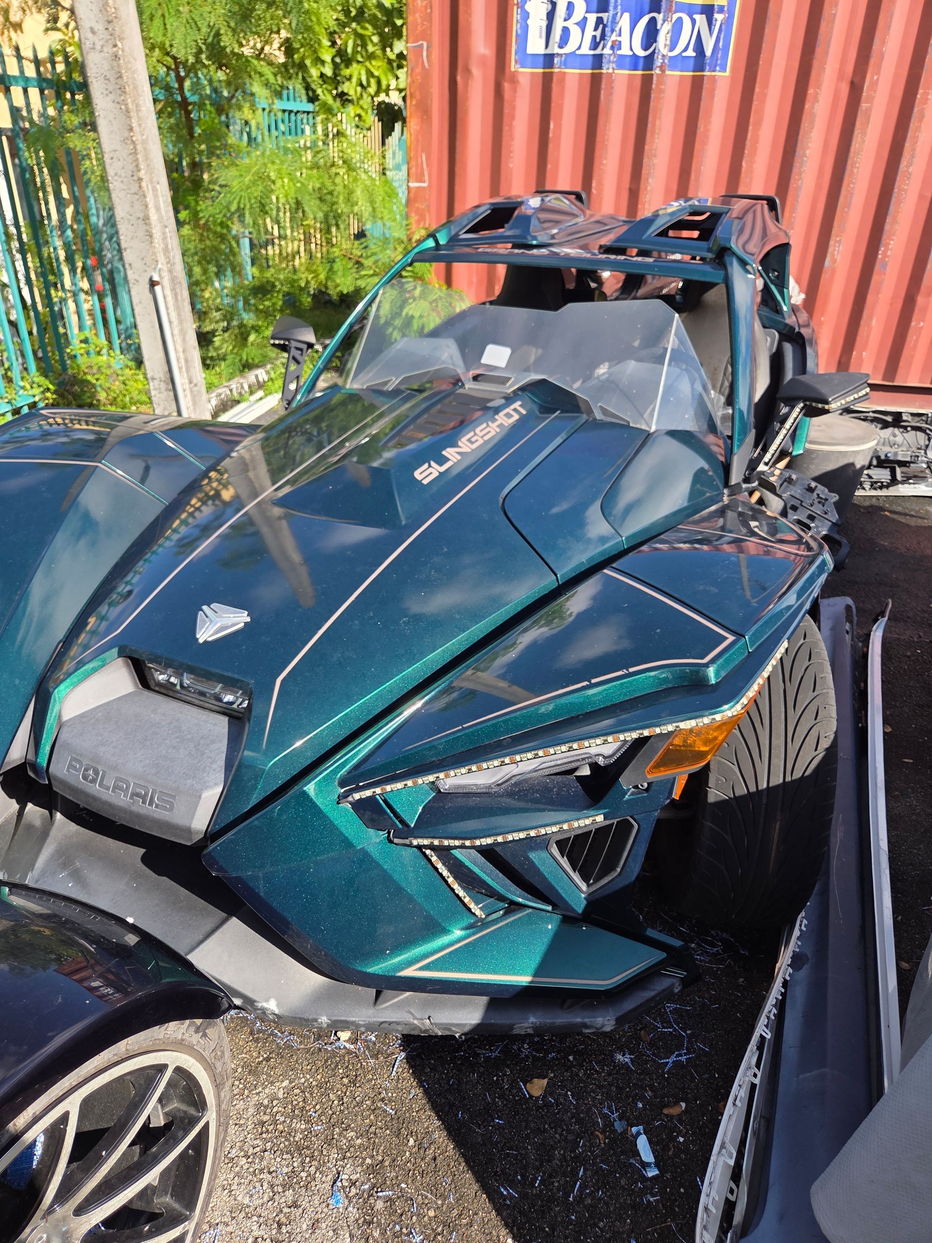 Damaged teal Polaris Slingshot three-wheeled vehicle after a crash. Debris is scattered.