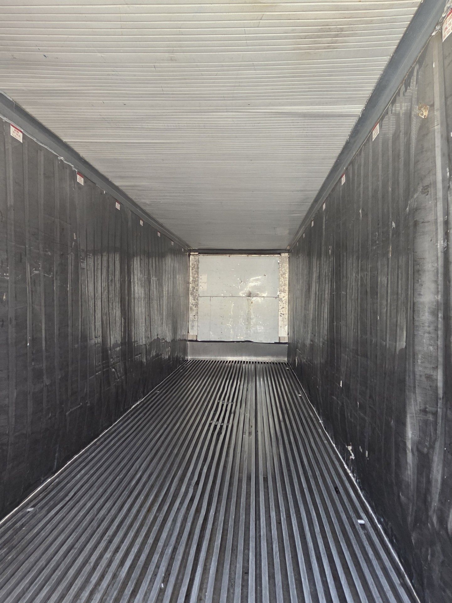 Empty cargo container interior, with ribbed metal floor and ceiling, walls in shadow, door at far end.