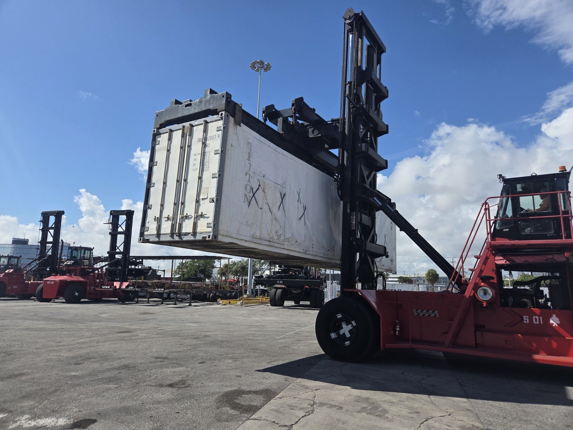 Red forklift lifting a large, white shipping container at an outdoor industrial site, with blue sky.