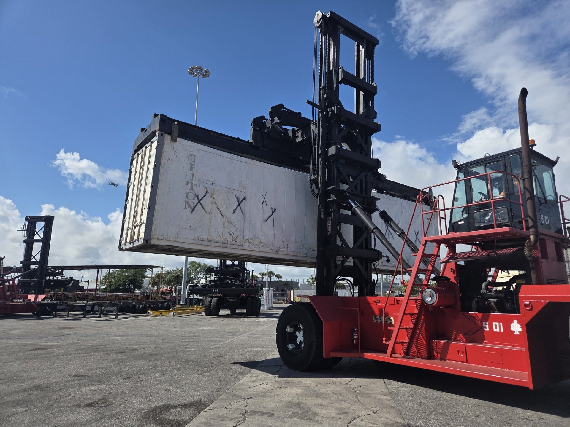 Red forklift lifting a large white shipping container at a port under a blue sky.