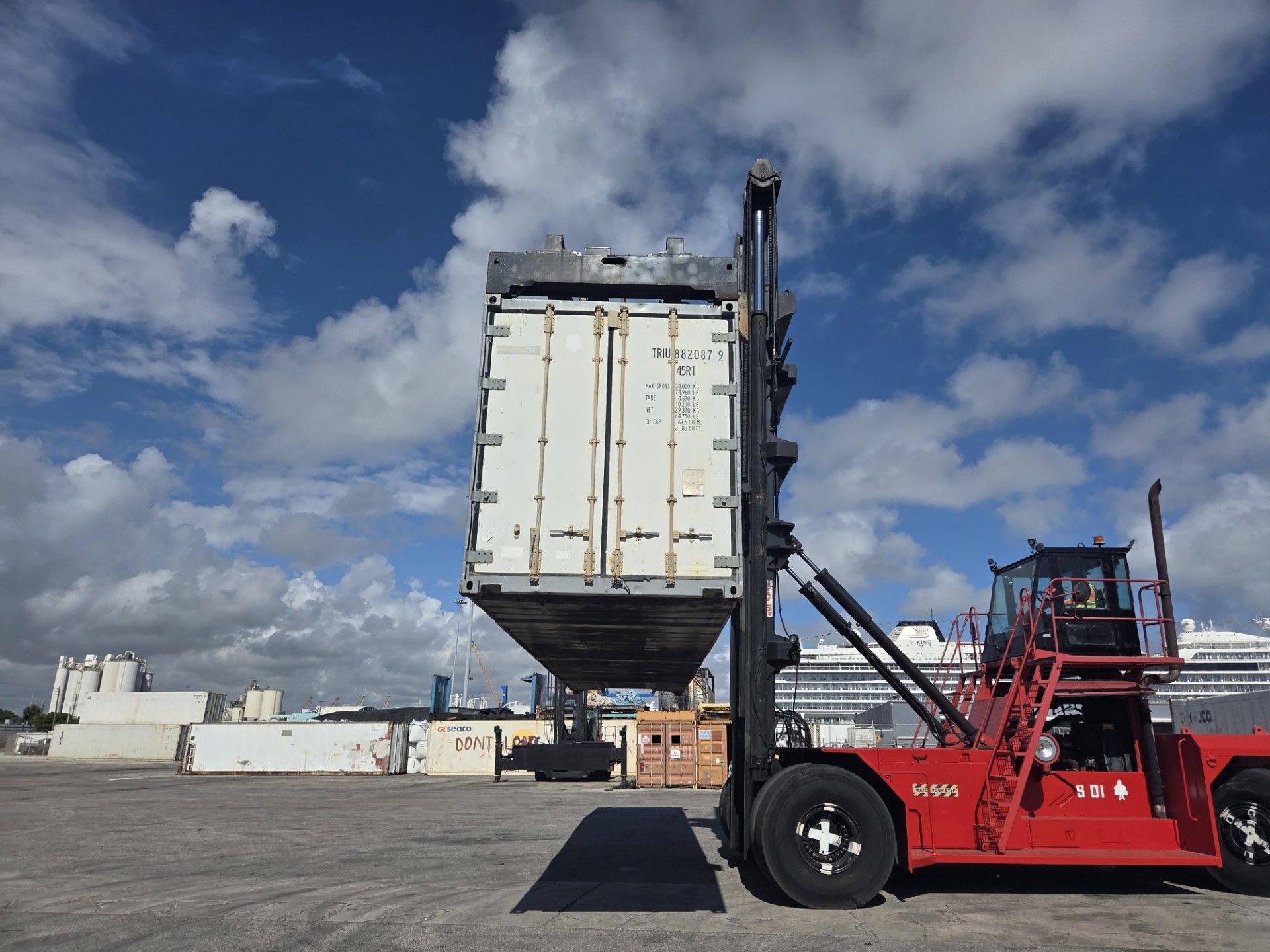 Red forklift lifting a white shipping container in a port on a sunny day with clouds.