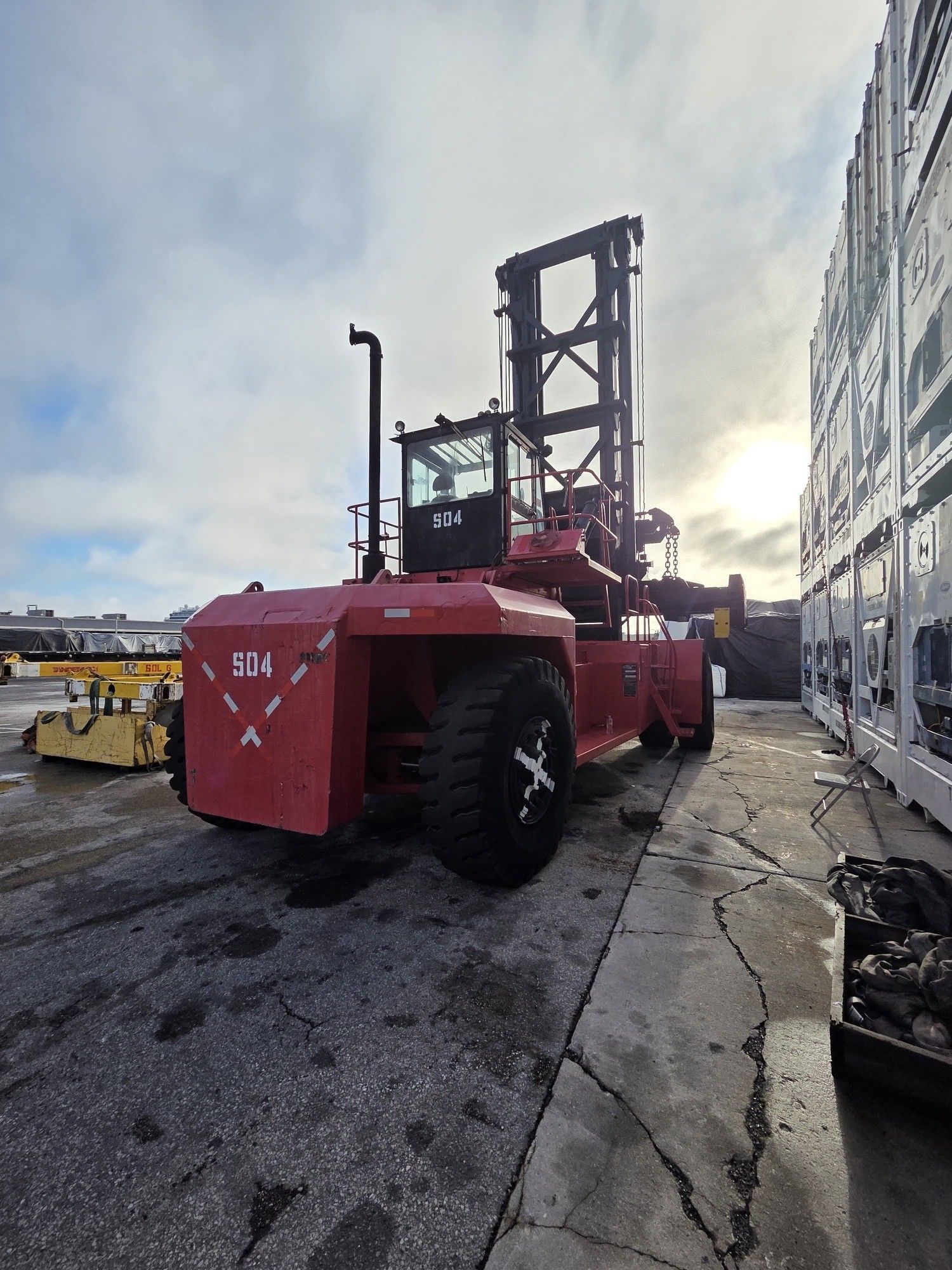 Red container handler machine in an industrial setting, next to cargo containers, under a cloudy sky.