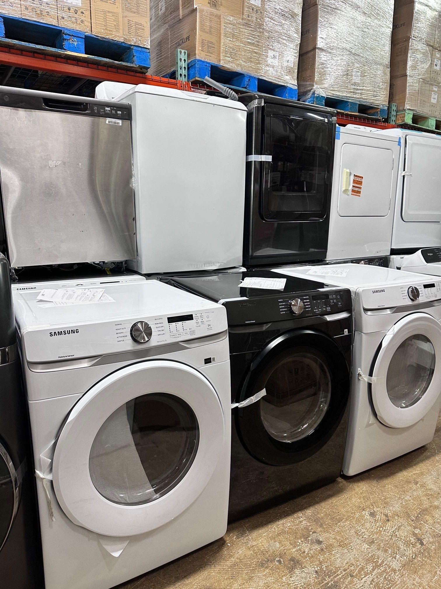 Row of washers and dryers, various colors including white, black, and stainless steel, in a warehouse setting.