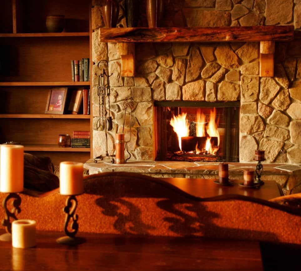 Cozy living room with stone fireplace, burning fire, wooden mantel, and bookshelves. Warm lighting and candles.