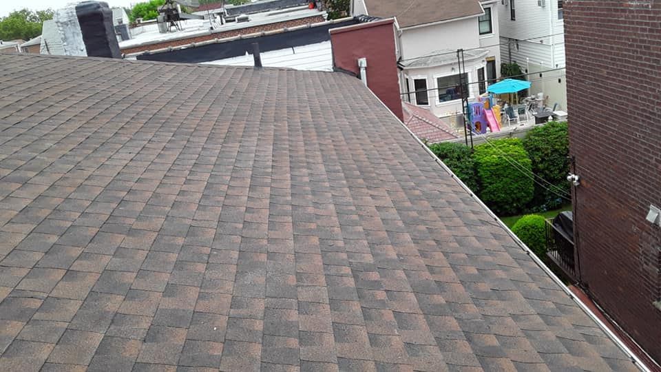 Brown asphalt shingle roof on a city building with other buildings and a backyard in the background.