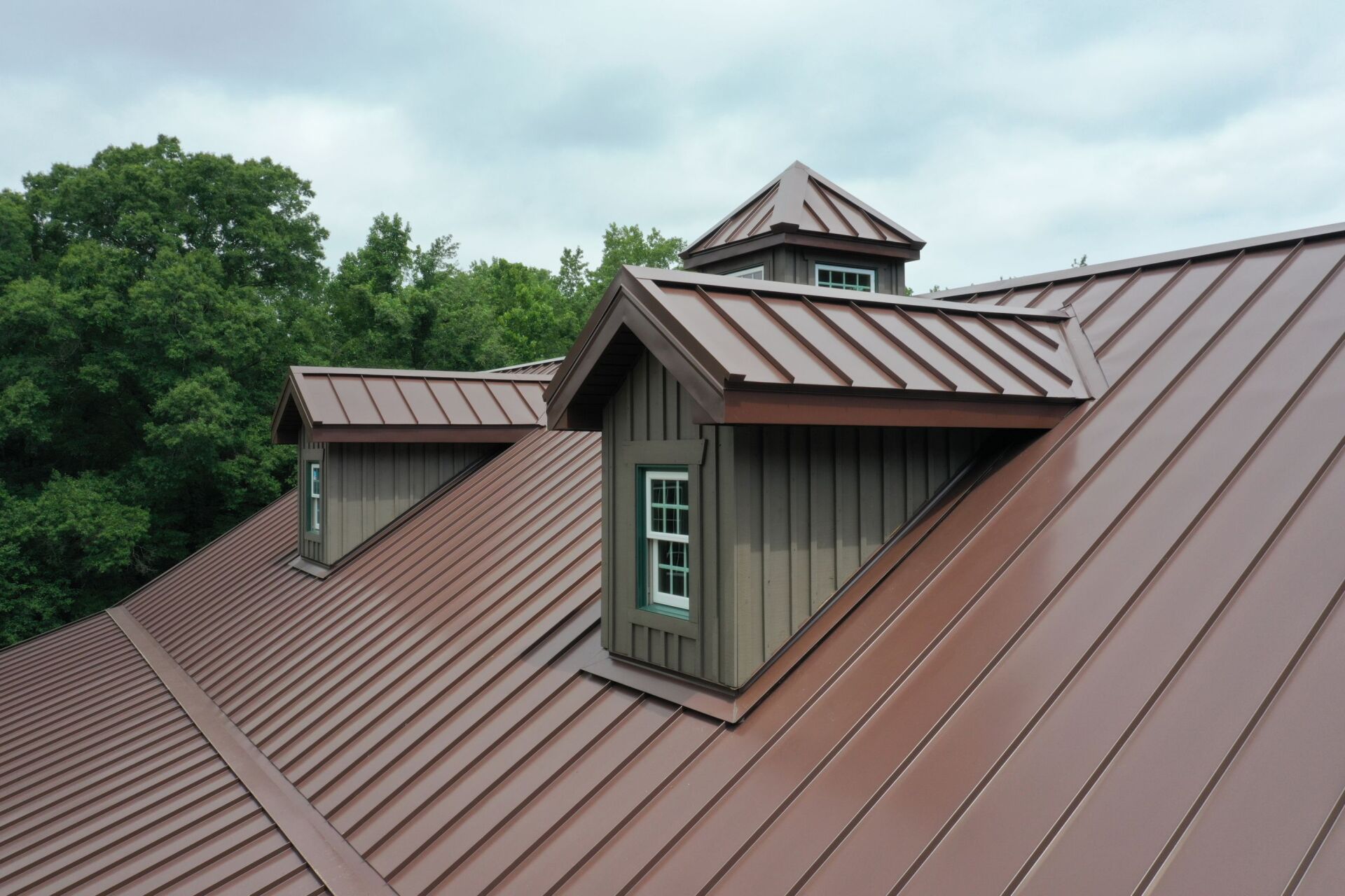 Brown metal roof with dormers, set against a green tree-filled background and cloudy sky.