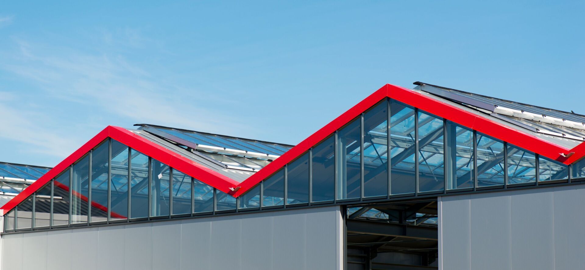 Red-roofed industrial building with sawtooth roof, clear glass panels, and blue sky background.