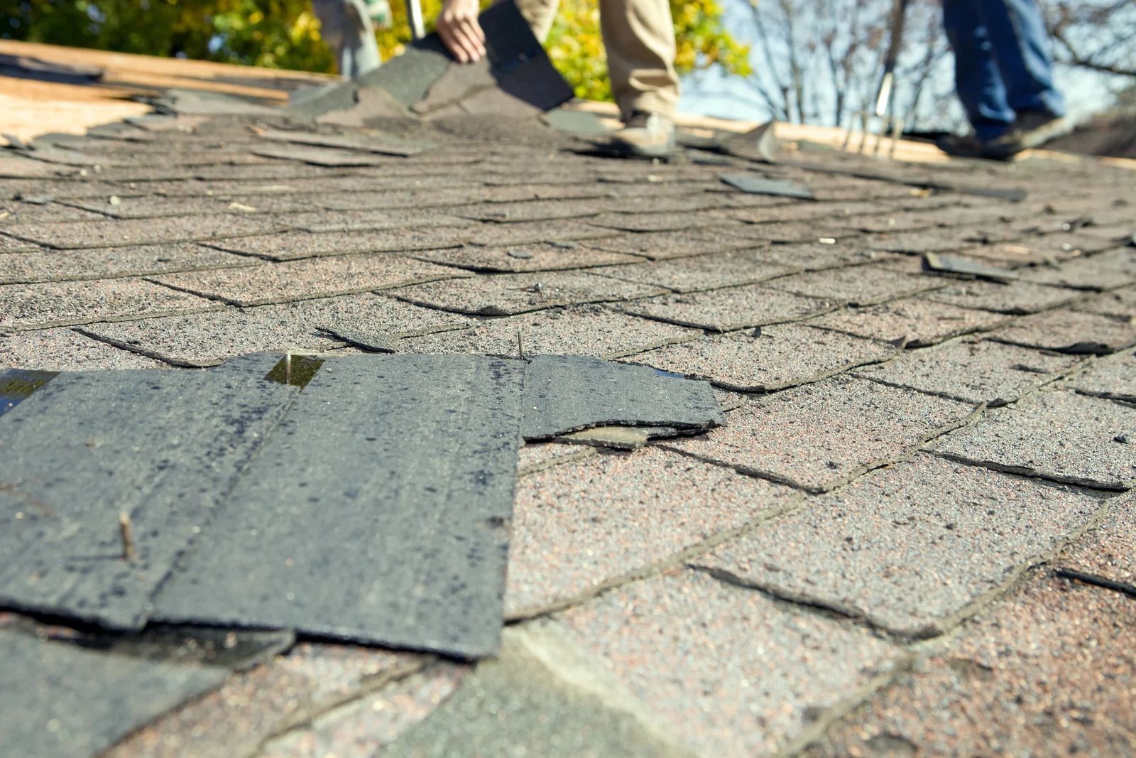Loose roof shingles on a rooftop with workers’ feet visible in the background