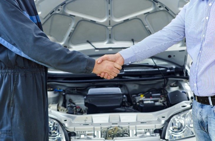 Two people shaking hands in front of an open car hood at a dealership