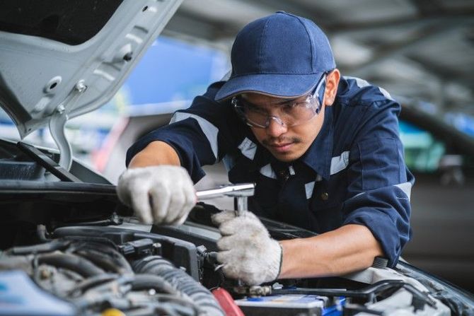 Mechanic in blue uniform using a wrench under an open car hood in a garage