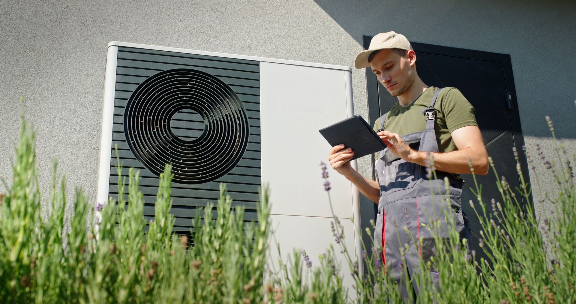 Technician using a tablet to inspect an outdoor heat pump unit. He wears a cap and overalls outside a house.