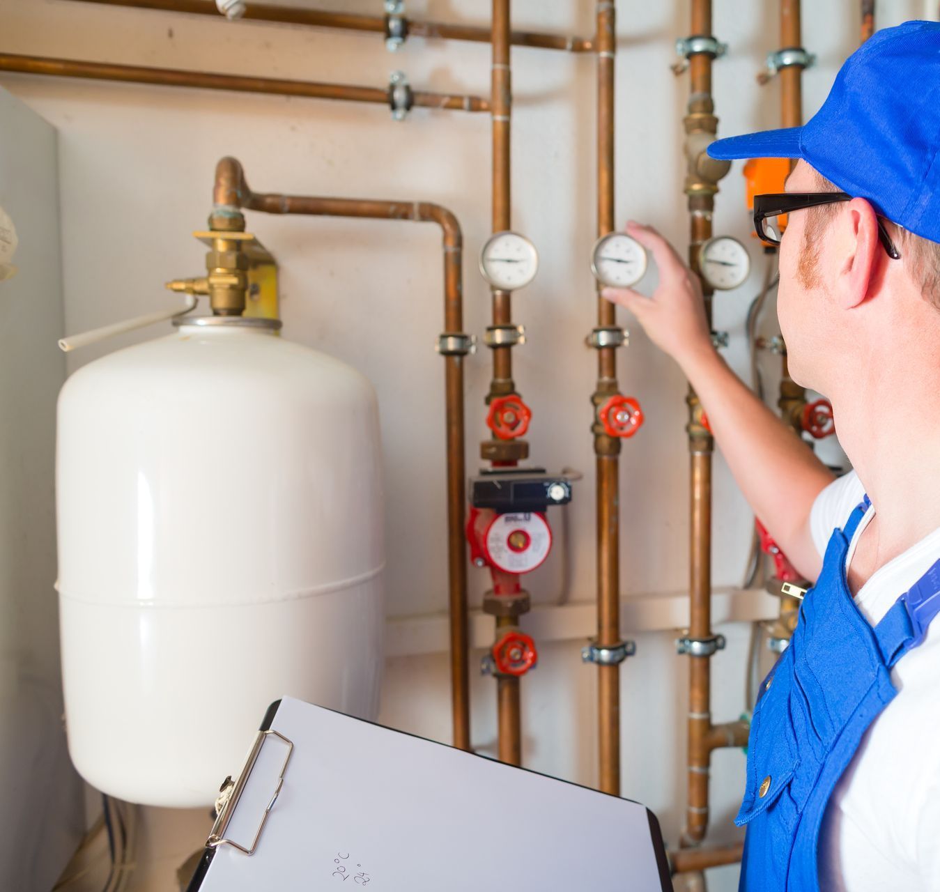 A plumber is working on a pipe in a room while holding a clipboard.