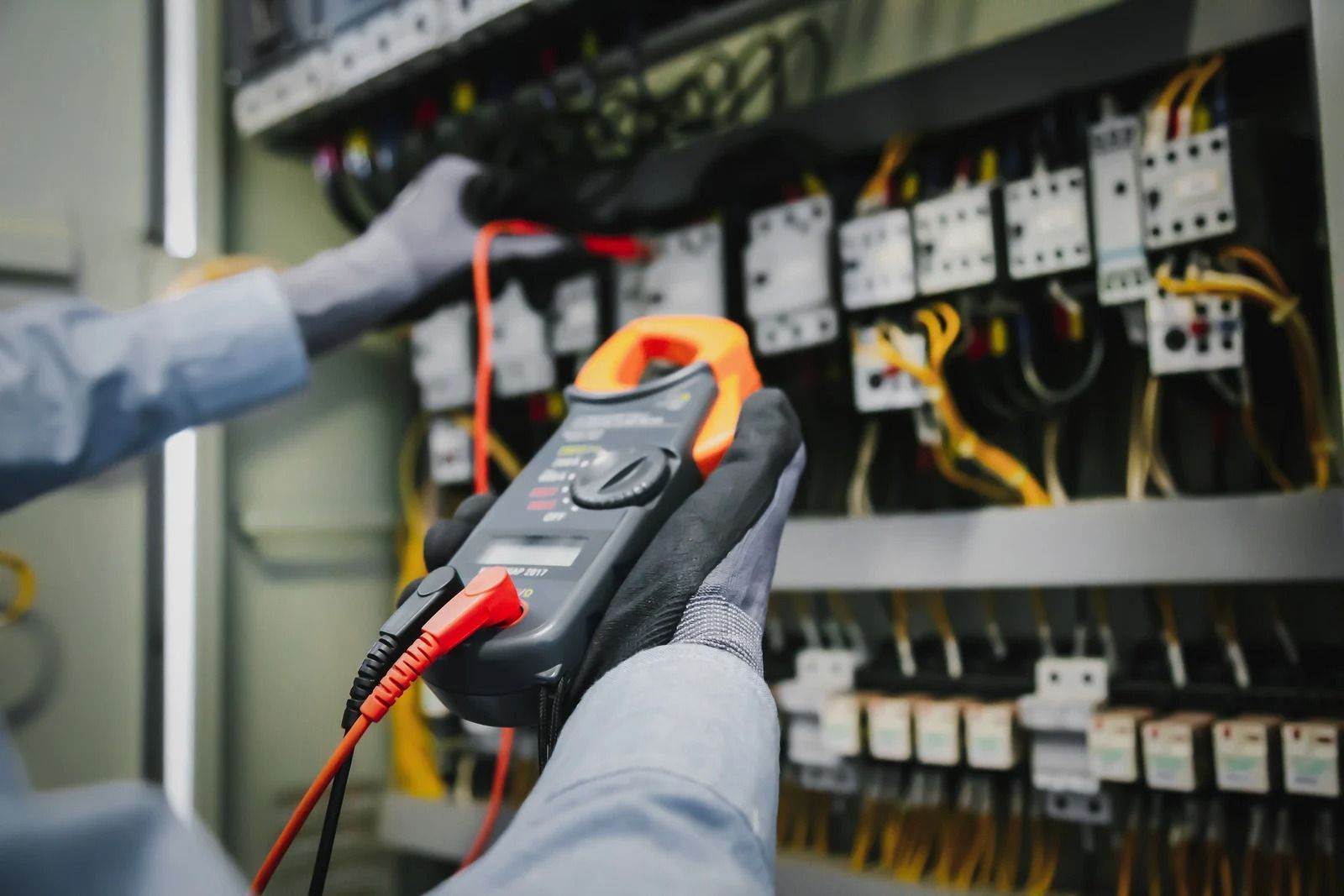 A worker in gloves uses a digital clamp meter to test electrical components in a control panel.