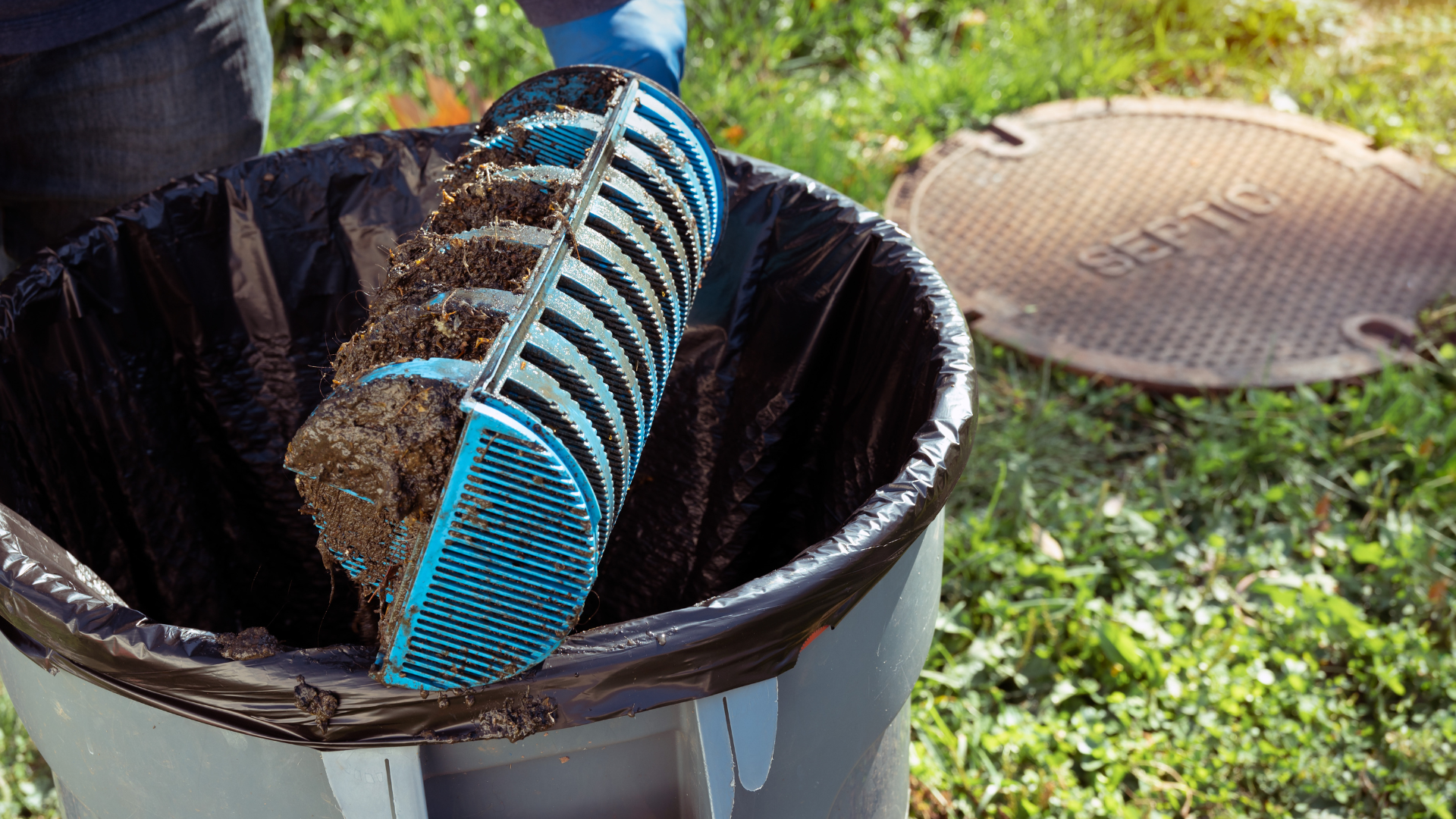 A person emptying a filter filled with debris into a black trash bag near a septic tank cover.