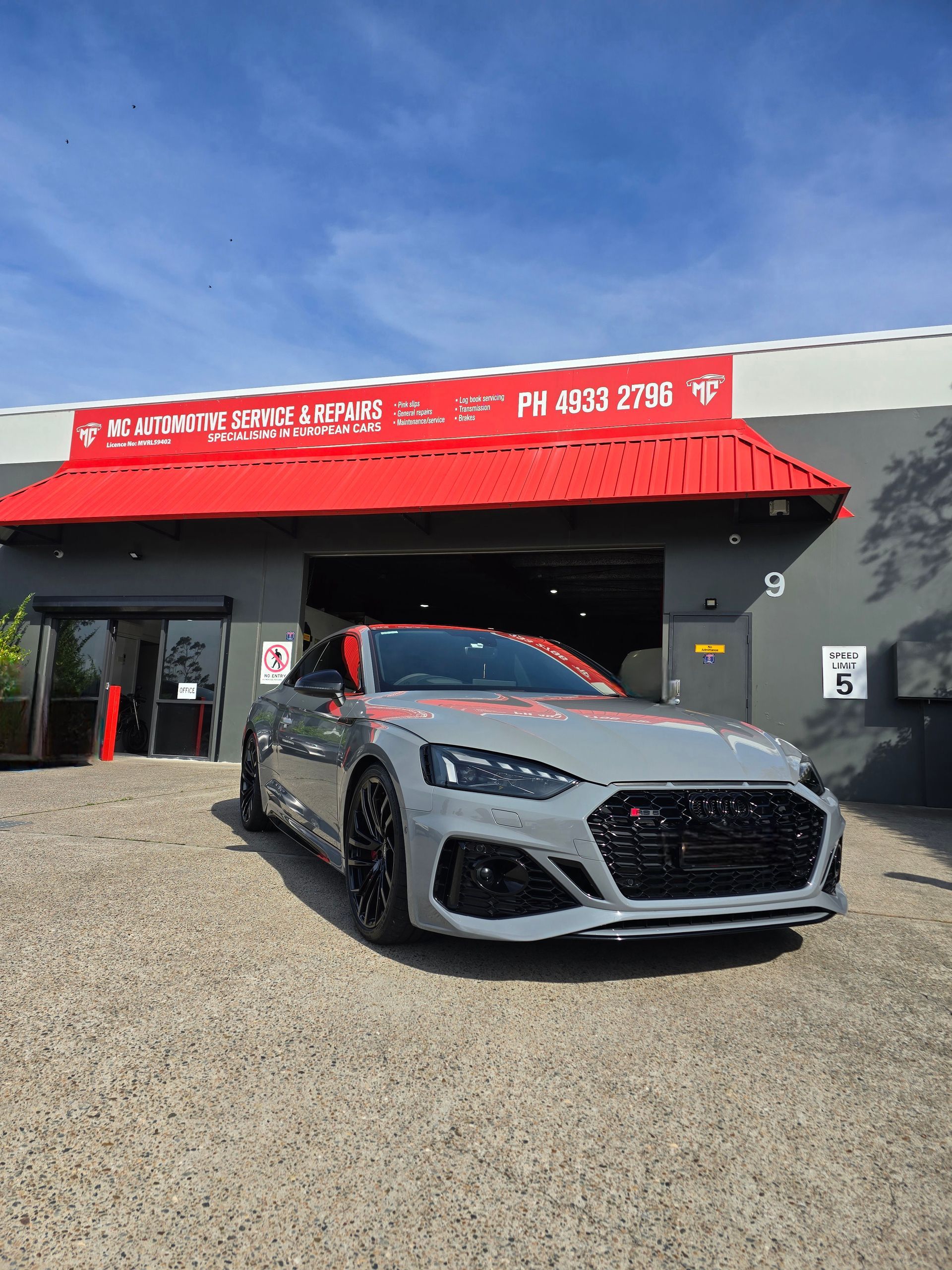 Gray sports car parked in front of a repair shop with red accents under a blue sky.