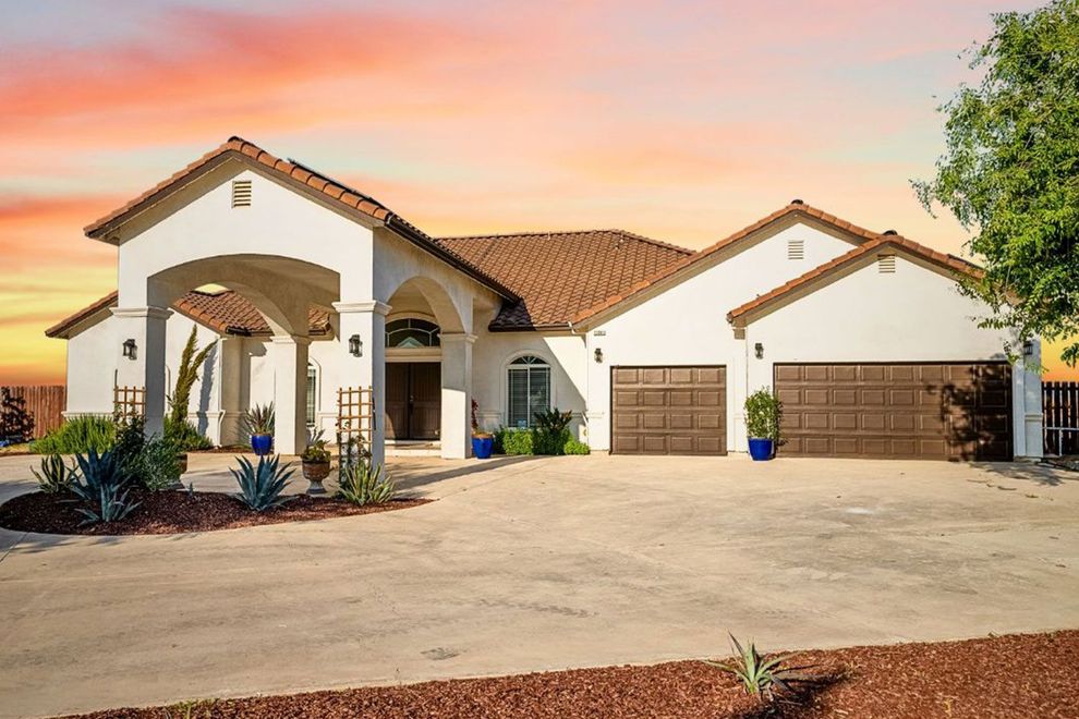 A stucco house with a terracotta roof, arched entrance, and a circular driveway under an orange sky.