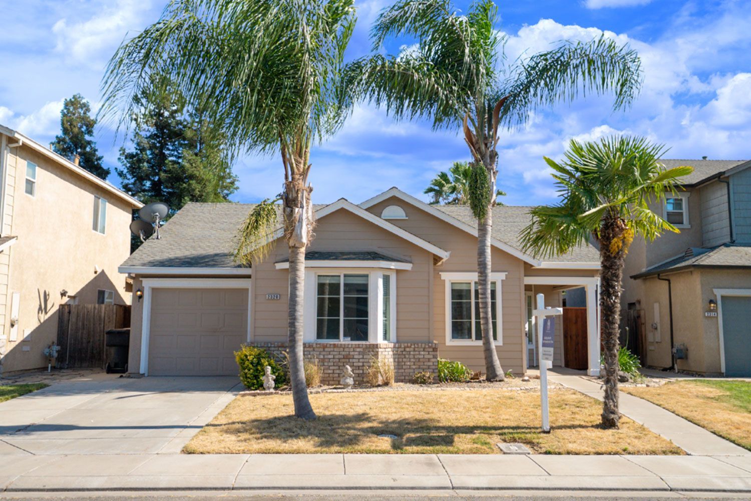 Tan suburban house with palm trees, blue sky, and a for sale sign.