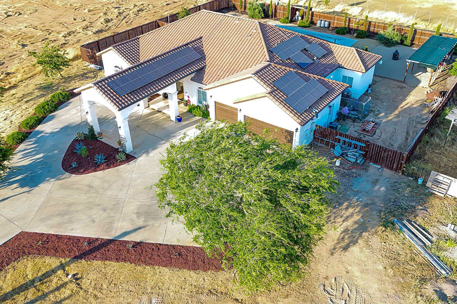House with solar panels on the roof and a large tree in front of a driveway, set in a dry, rural landscape.