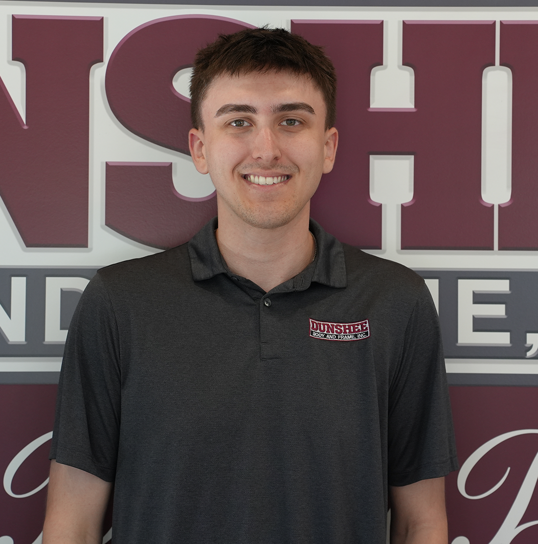 Smiling person in a dark polo shirt standing before a maroon and gray NSH sign
