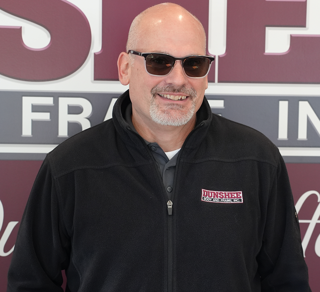 Bald man in sunglasses and black zip-up jacket standing in front of a store sign