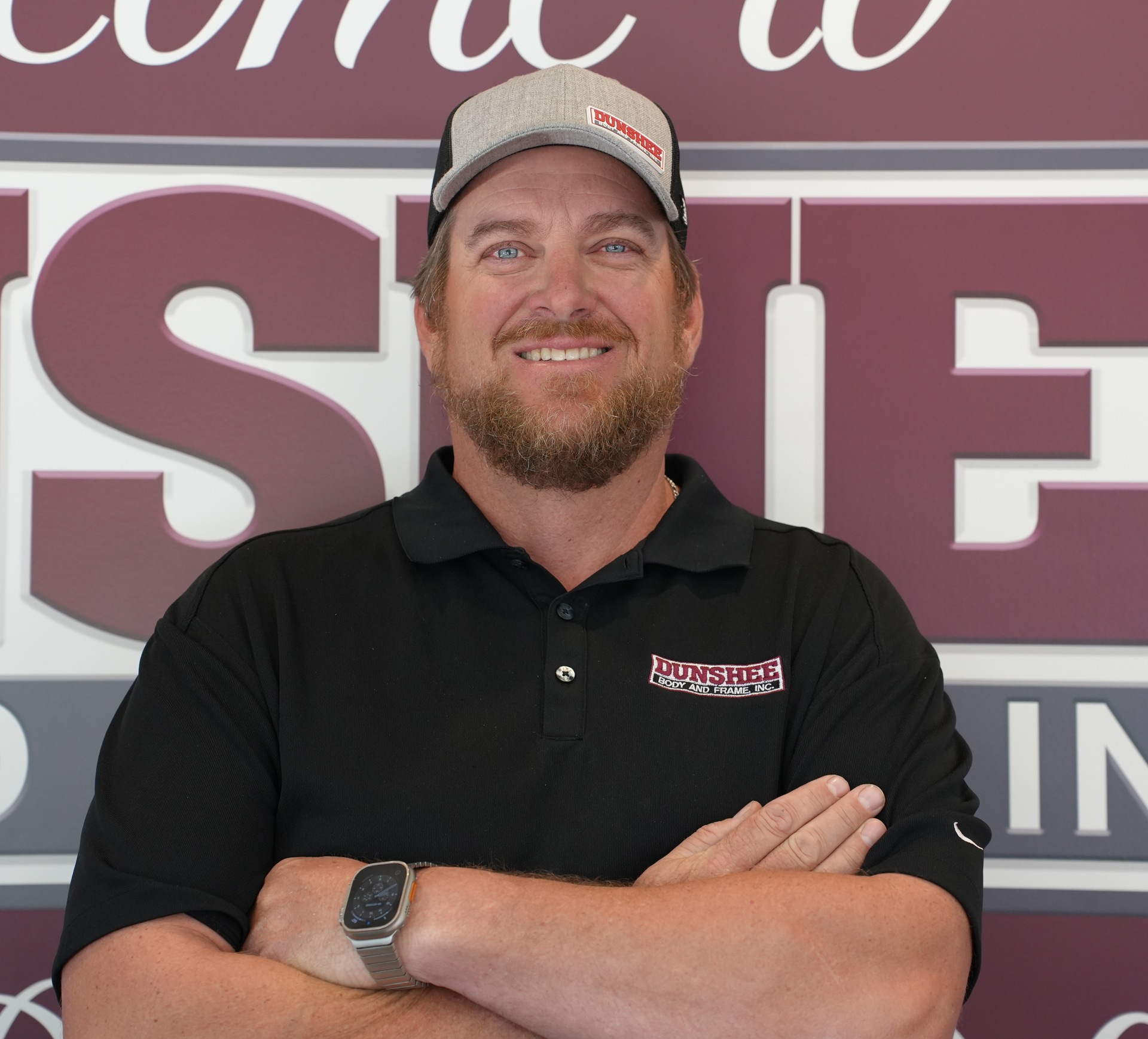 Smiling man in black polo and cap with arms crossed in front of a sign