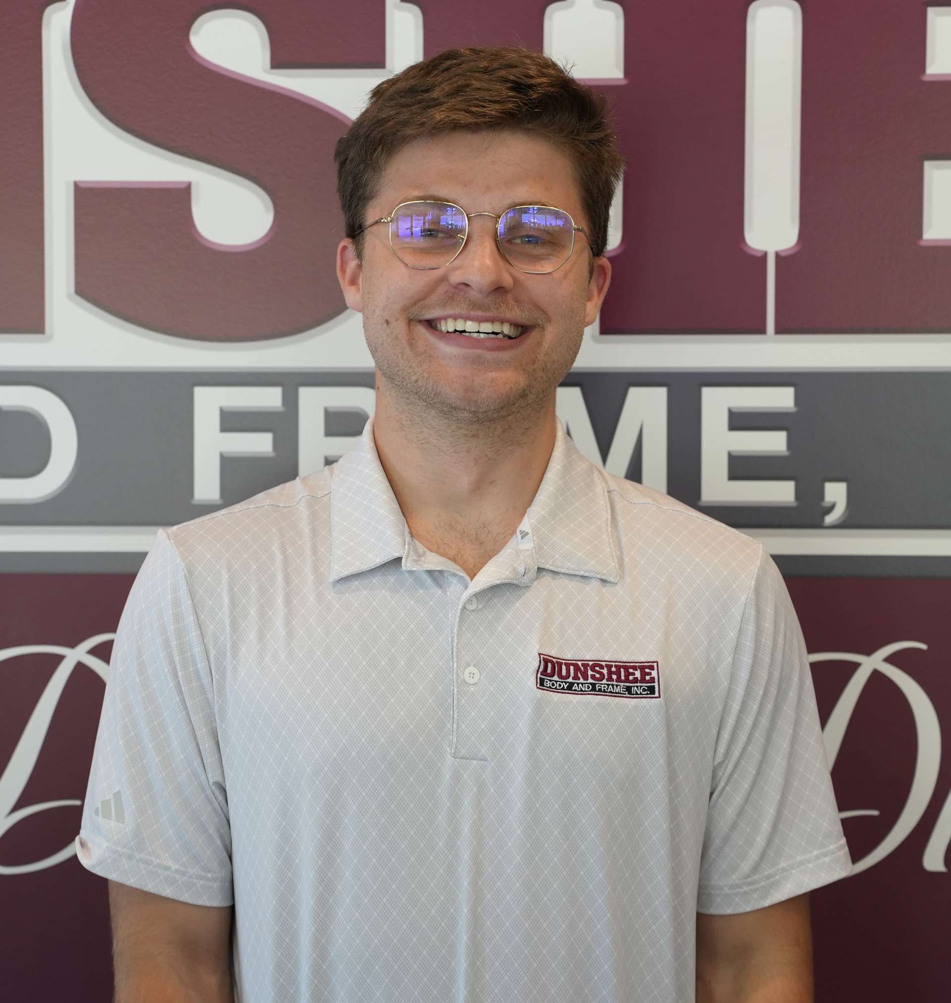 Smiling man in glasses wearing a white polo shirt stands before a maroon and gray wall sign.