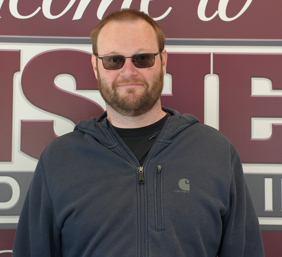 Man wearing sunglasses and a navy jacket standing before a maroon-and-white sign