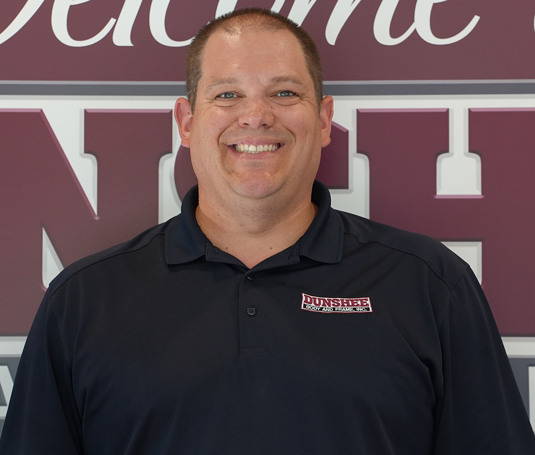 Smiling man in a black polo shirt standing in front of a maroon and white sign.