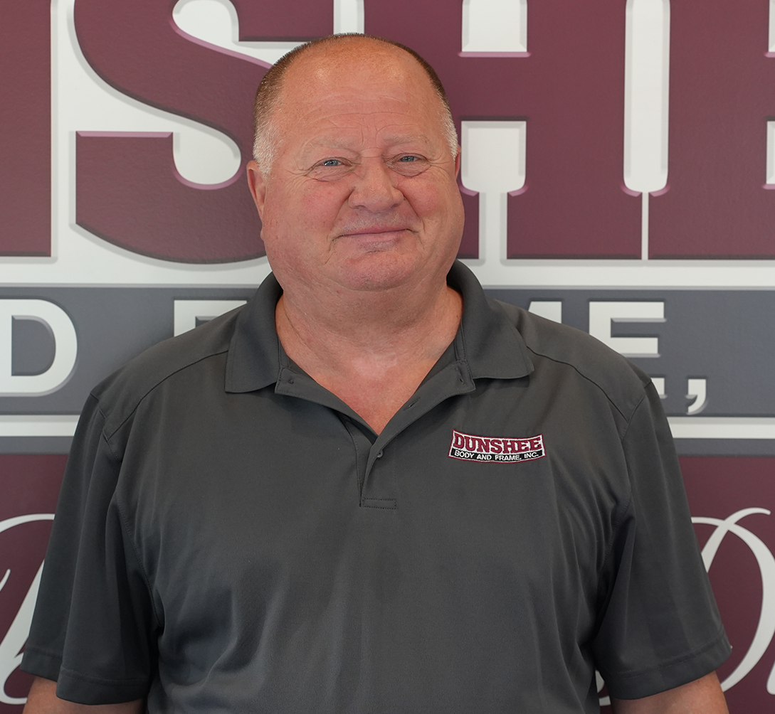Man in a dark gray polo shirt standing in front of a maroon and gray sign, smiling gently.