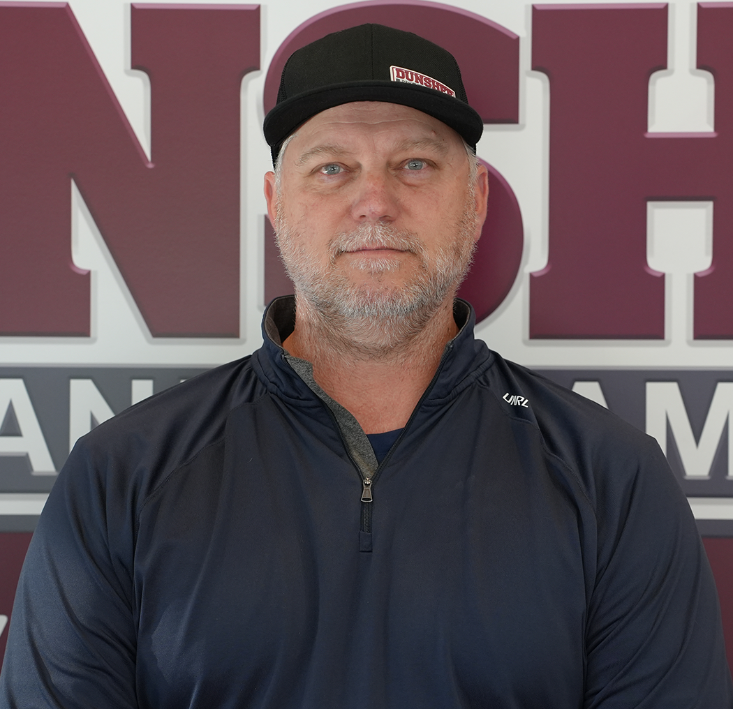 Man in a black cap and jacket standing before a maroon sign with white lettering