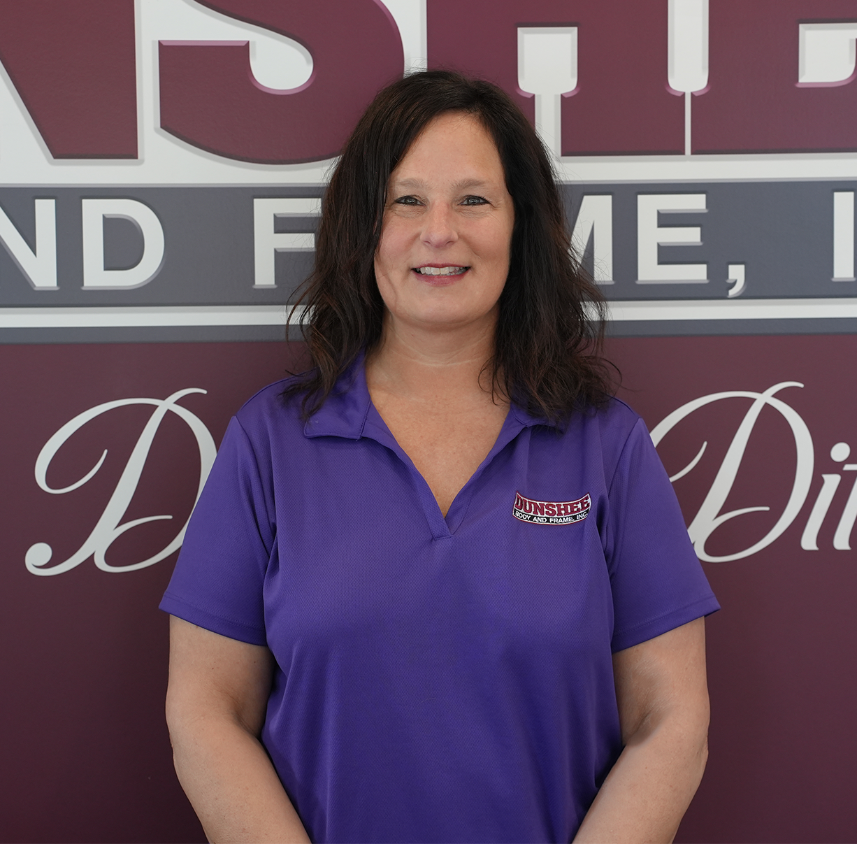 Woman in a purple polo shirt standing in front of a sign, smiling at the camera
