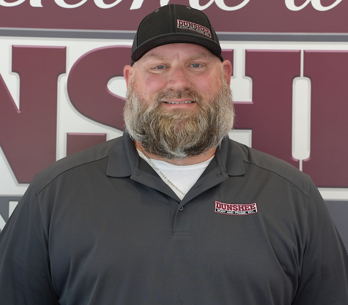 Smiling bearded man in a black cap and polo shirt standing before a red and white sign