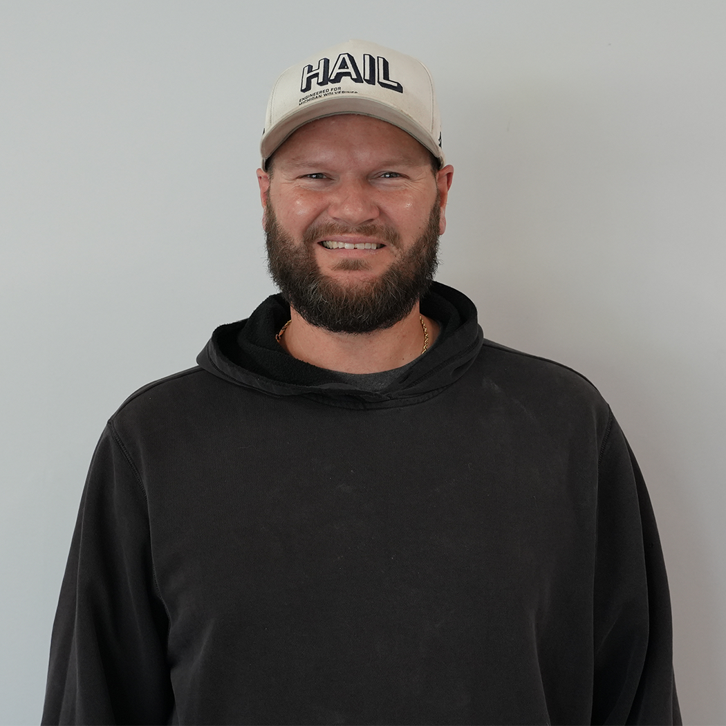 Smiling person in a black hoodie and beige cap against a plain gray wall