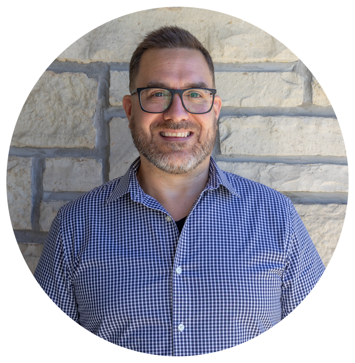 A man in a blue plaid shirt is smiling in front of a brick wall