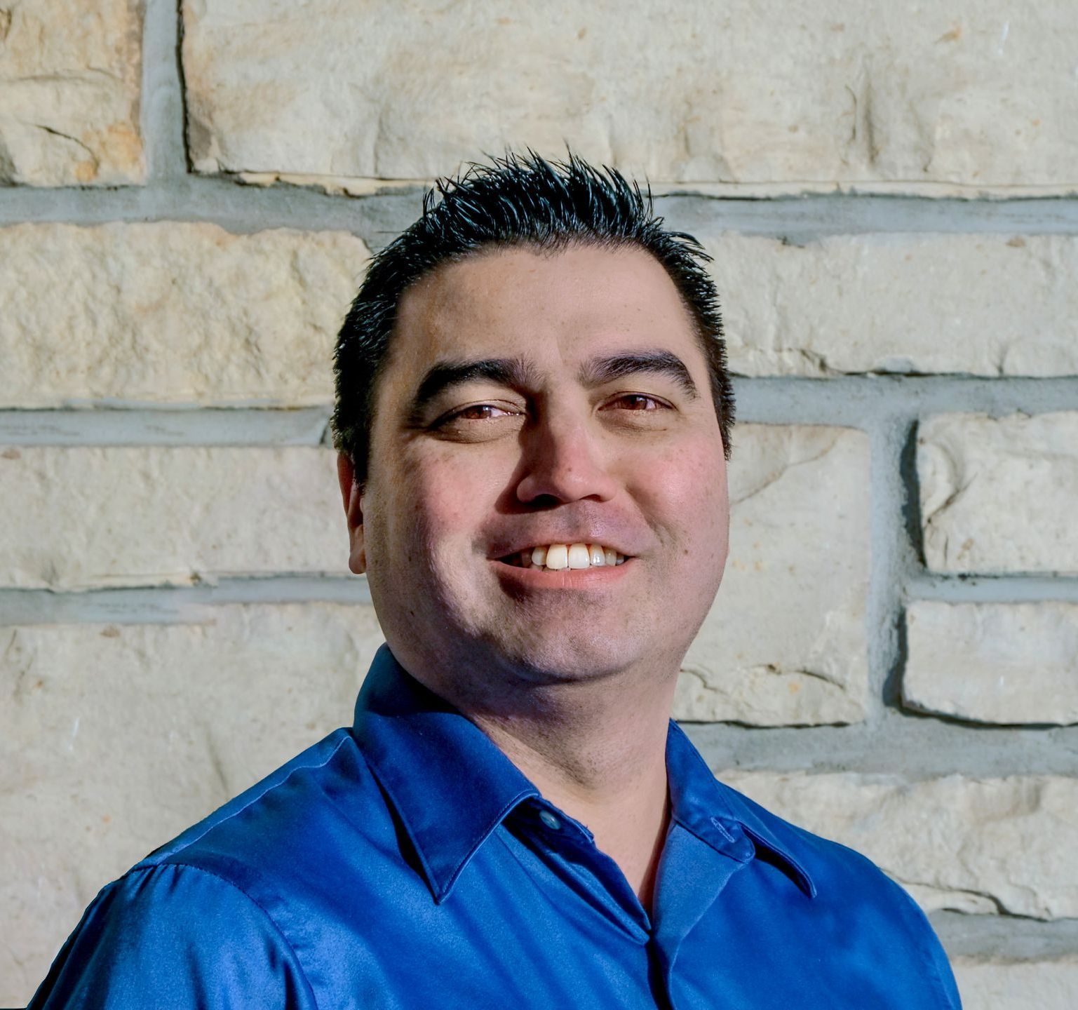 A man in a blue shirt is smiling in front of a brick wall